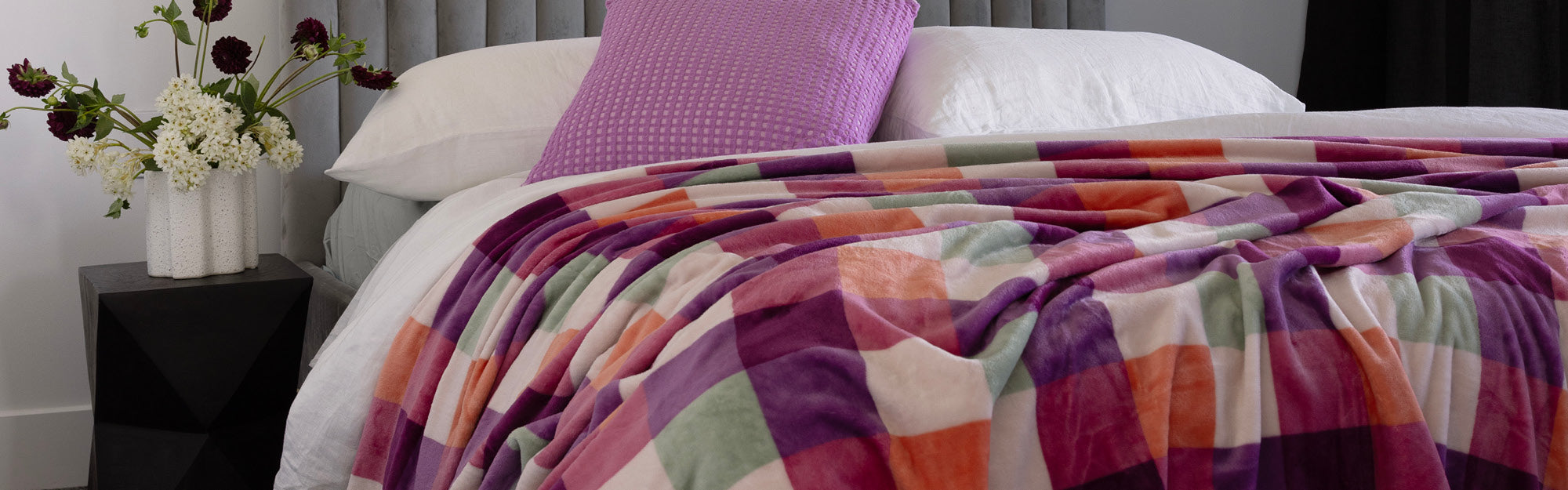 Bambury's Charlie Ultraplush colourful checkered blanket on a bed with a vase of flowers and a Orchid pink Dora cushion.